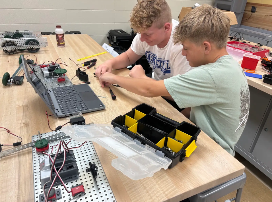 Two students sitting at a desk working on a robot with a laptop sitting in front of them