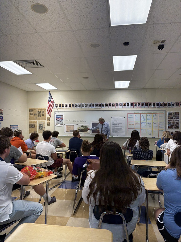 Students seated in chairs with their backs to the camera. A man presents from the front of the room. 