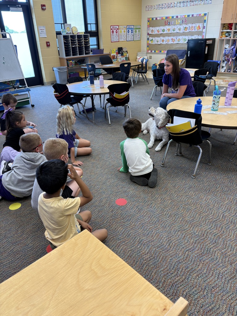 Students sitting on the carpet listing to an adult talk with a dog at her feet