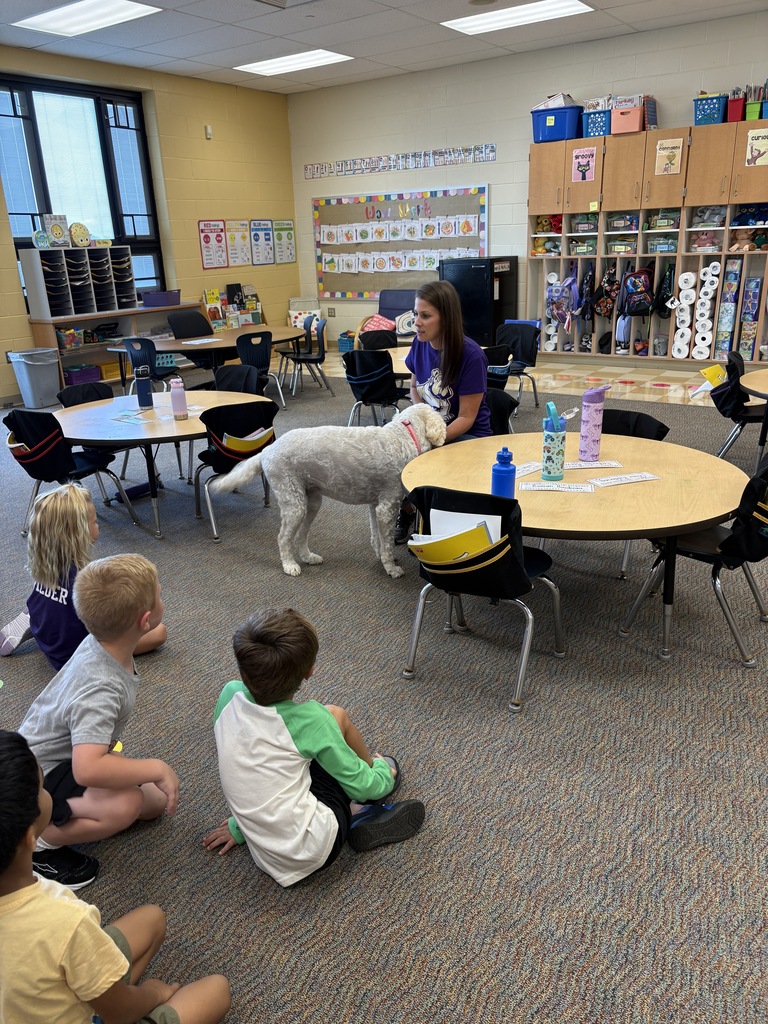 Students sitting on the carpet listing to an adult talk with a dog at her feet