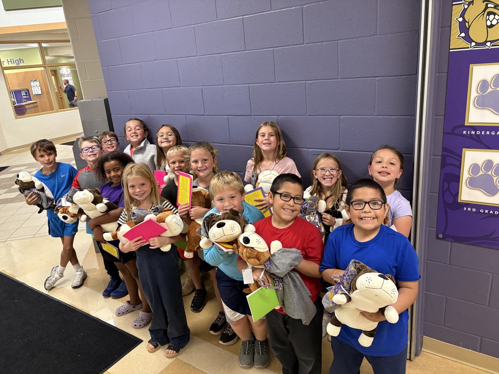 Young students standing in front of a purple wall holding stuffed dogs