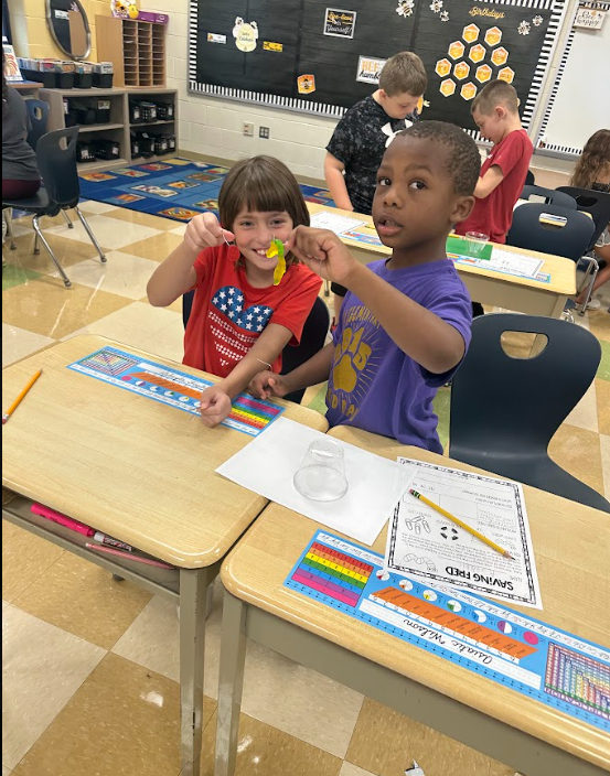 Two students using a clear plastic cup and a gummy worm standing at a desk