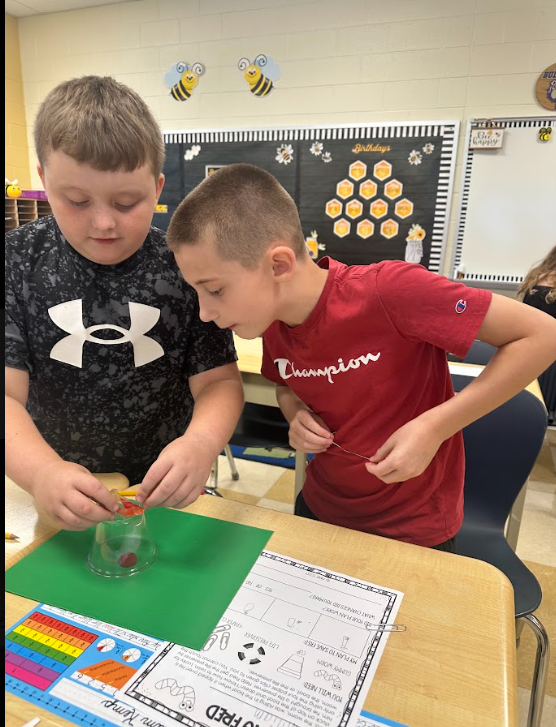 Two students using a clear plastic cup and a gummy worm standing at a desk