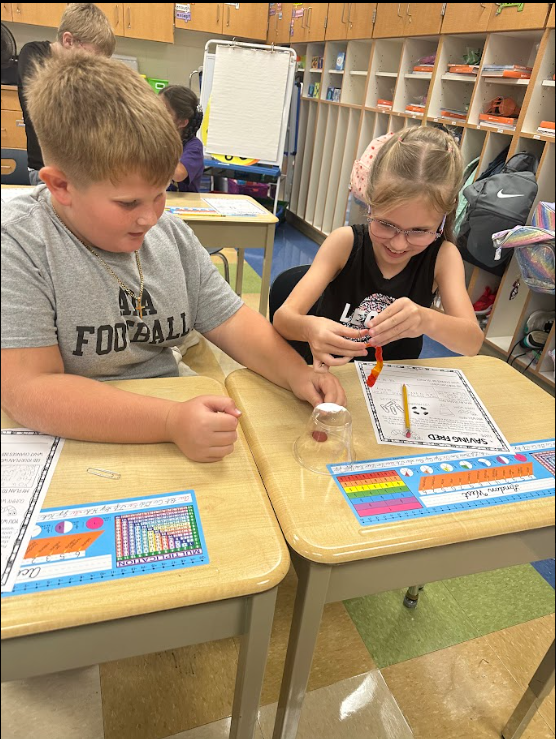 Two students using a clear plastic cup and a gummy worm standing at a desk