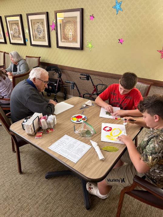 middle school students sitting at a table with elderly nursing home residents painting pictures