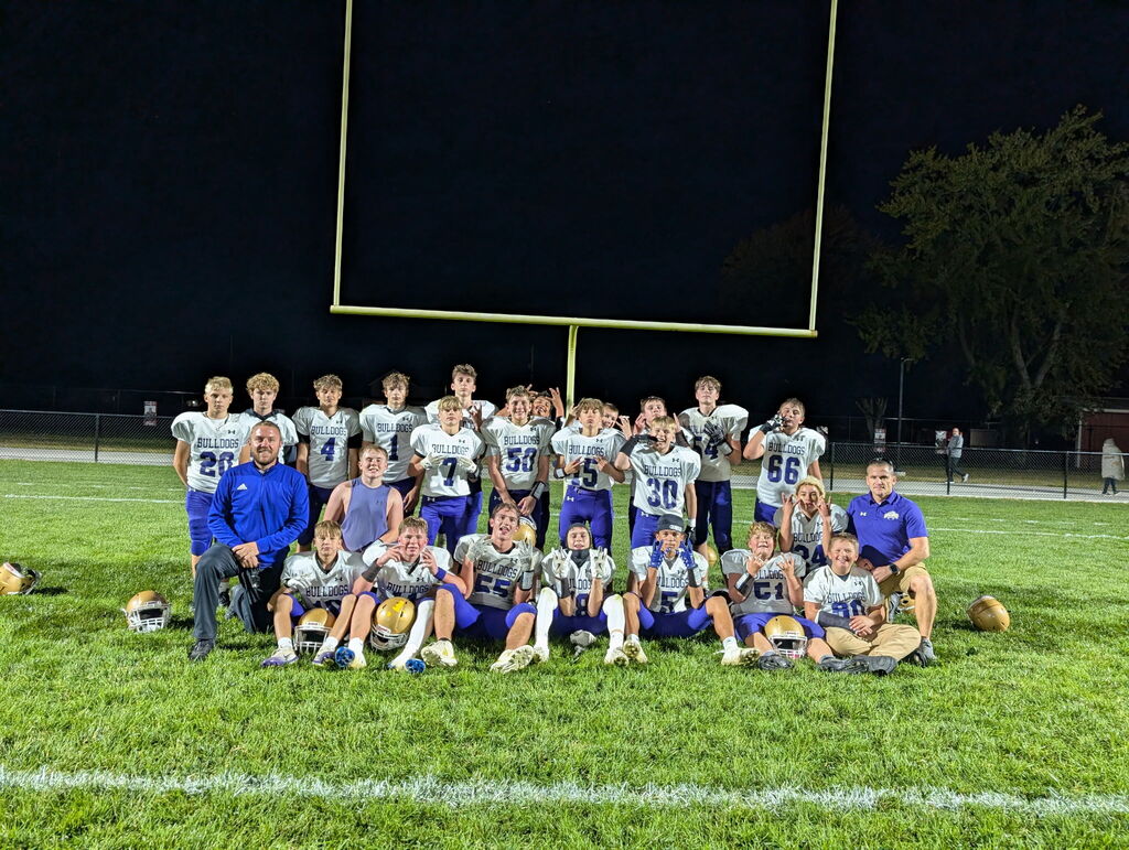 Young football players standing in front of the goal post posing for a photo with two coaches on each side of the group