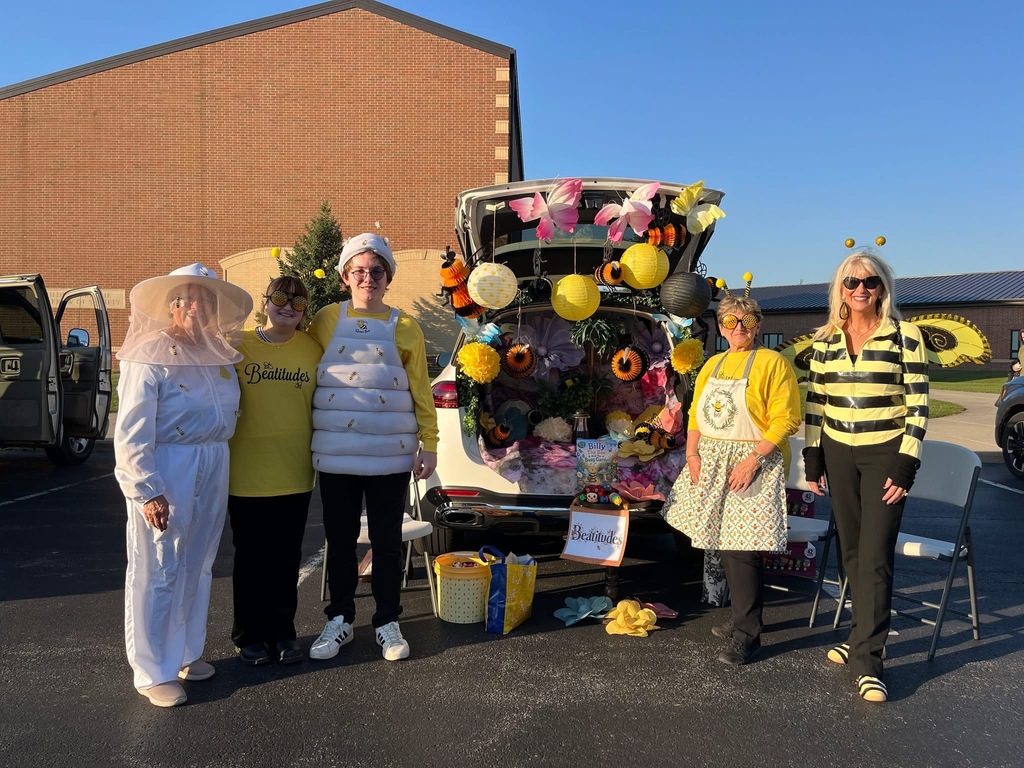 Five people dressed in white, yellow and bee outfits in front of an open trunk 