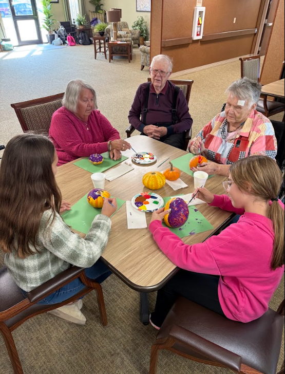 Three students sitting with elderly adults painting pumpkins around a table
