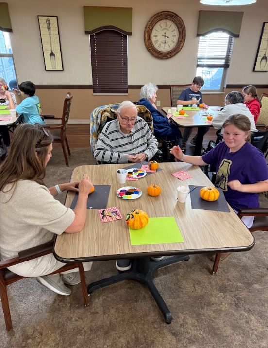 Students sitting with elderly adults painting pumpkins around a table