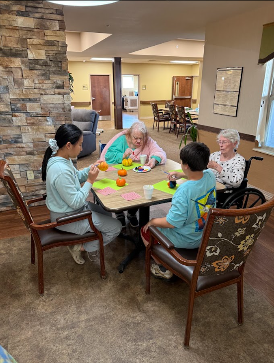 Two students sitting with elderly adults painting pumpkins around a table