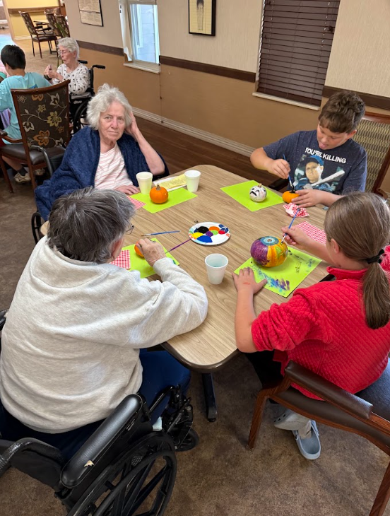 Twostudents sitting with elderly adults painting pumpkins around a table
