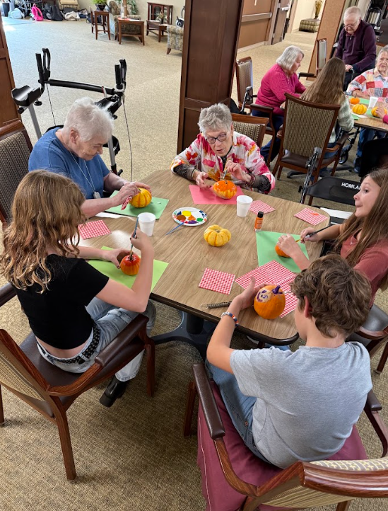 Three students sitting with elderly adults painting pumpkins around a table