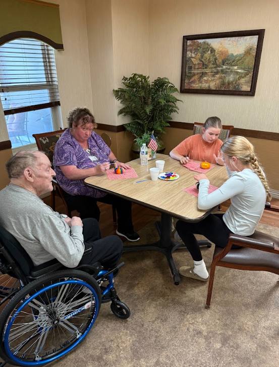 Two students sitting with elderly adults painting pumpkins around a table