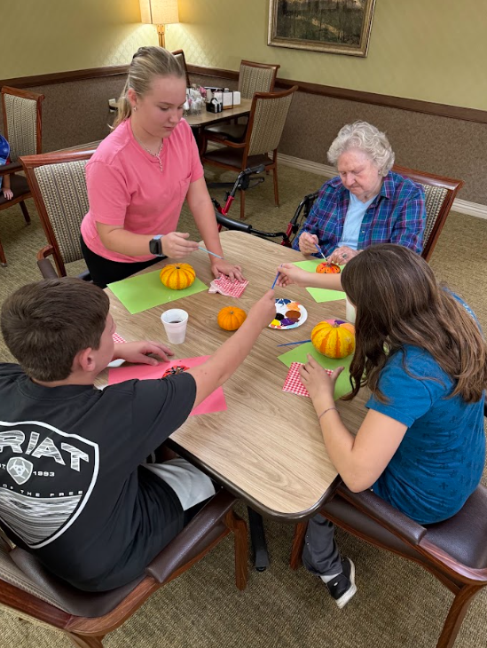 Three students sitting with elderly adults painting pumpkins around a table