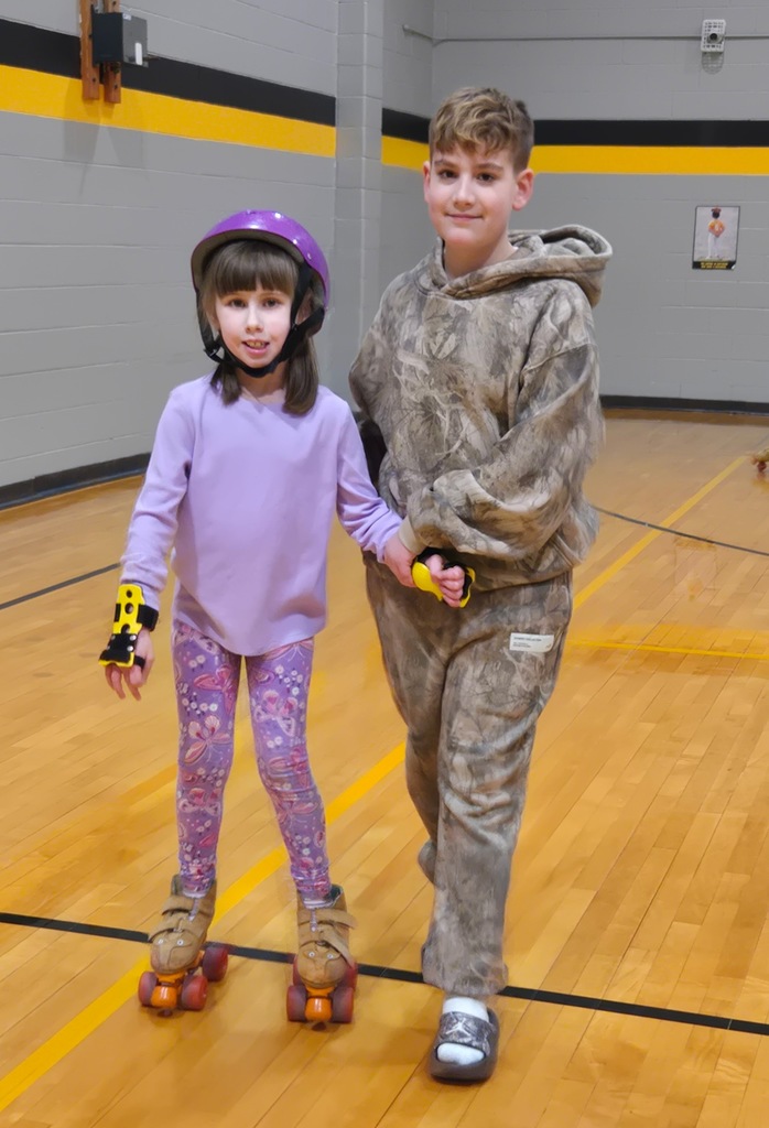 older male student holding hands with a smaller student helping her skate in the gym