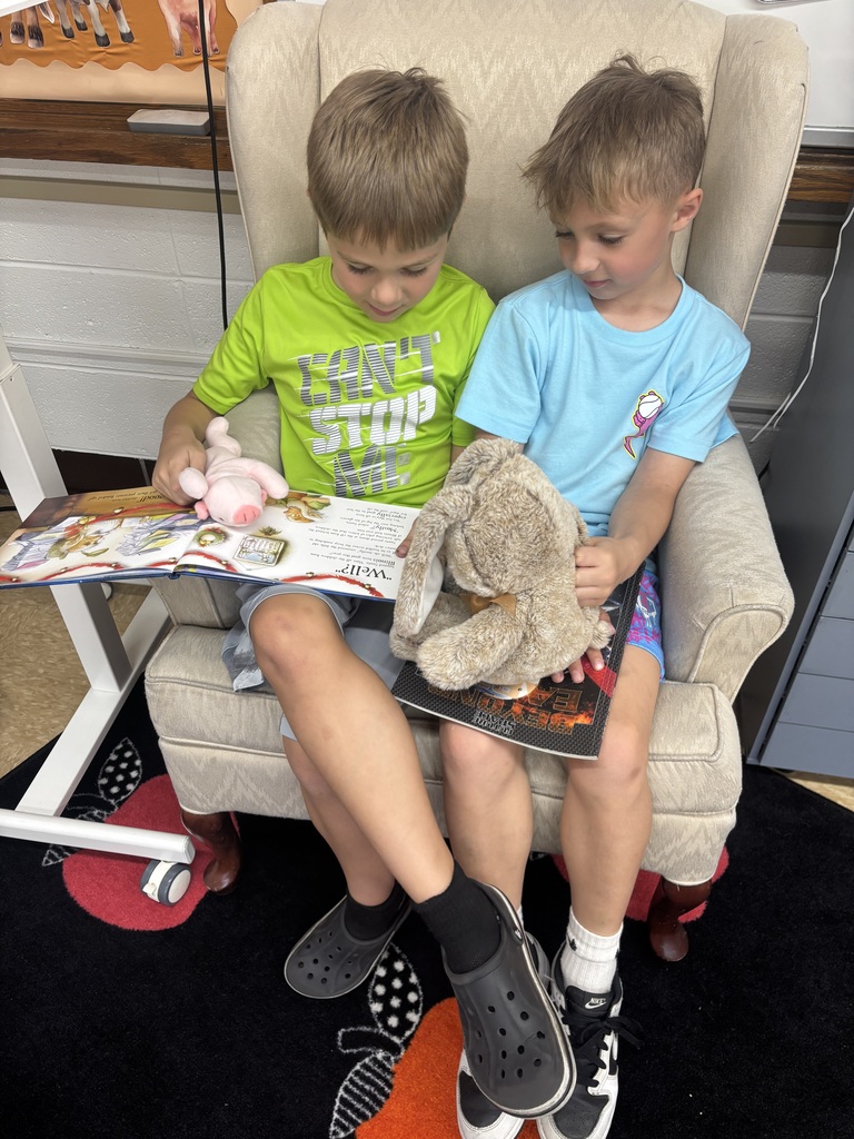 two male students sitting in a chair together reading with their stuffed animals