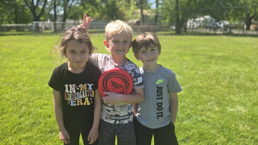 three students outside smiling holding a frisbee. Boy in middle giving bunny ears.