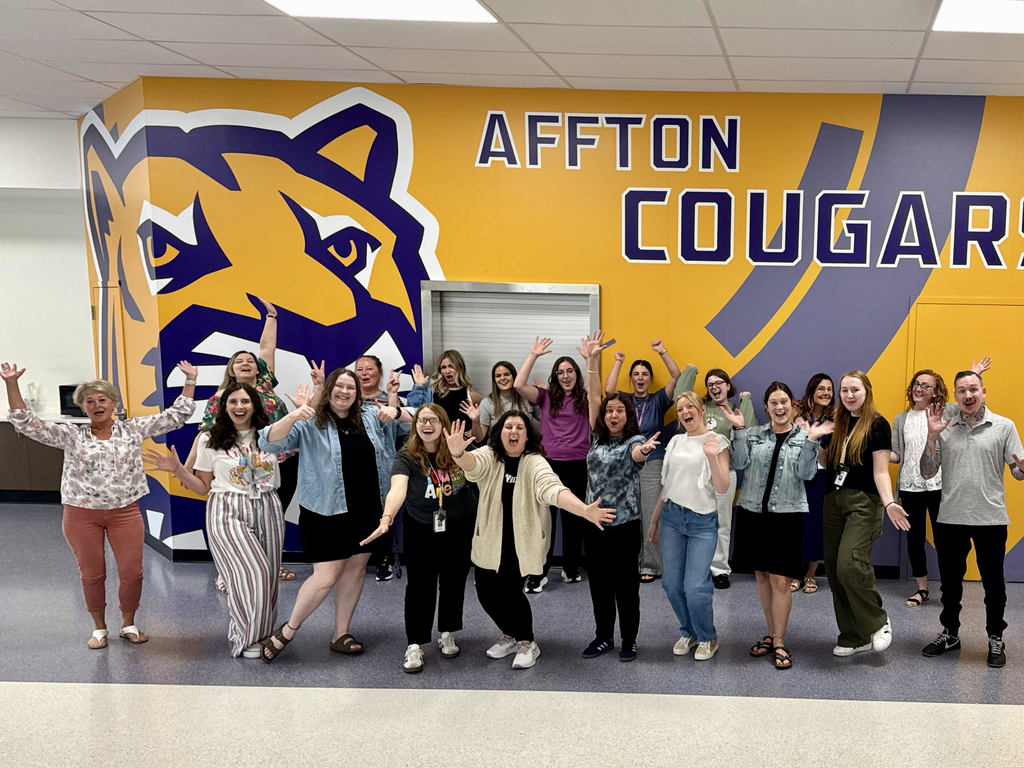 A group of 19 new teachers at Affton School District are posing for a lively photo in the high school cafeteria. They are all cheering and raising their hands in the air, with big smiles on their faces. The background is a bright yellow and purple wall with "AFFTON COUGARS" written in large letters and a graphic of a cougar head.