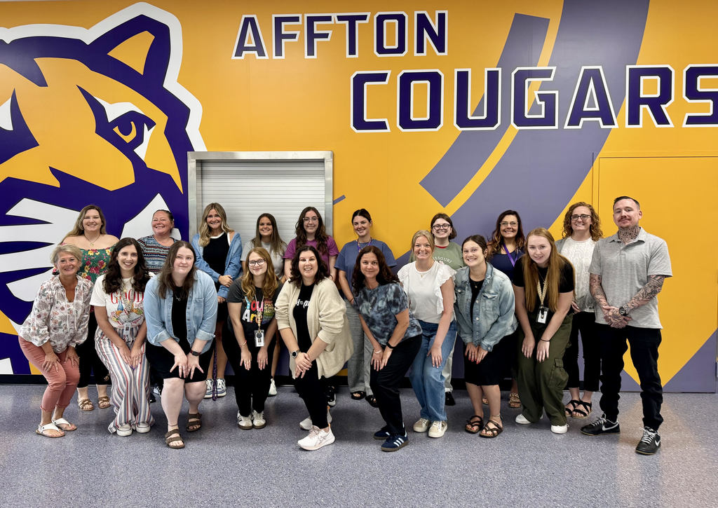 A group of 19 new teachers at Affton School District are posing for a photo in the high school cafeteria. They are a diverse group of adults, a mix of men and women, smiling and dressed casually. The photo's background is a bright yellow and purple wall with "AFFTON COUGARS" written in large letters and a graphic of a cougar head.