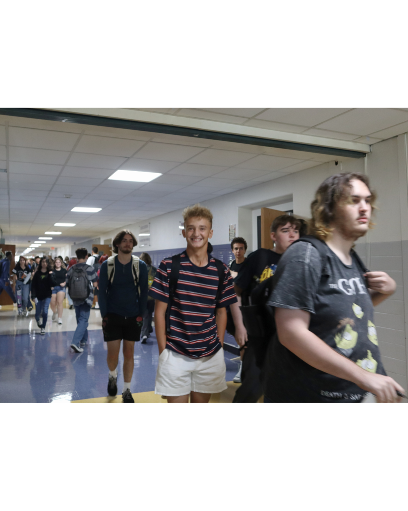High School students walk through the hallway; one high school boy in a striped shirt and shorts is smiling at the camera
