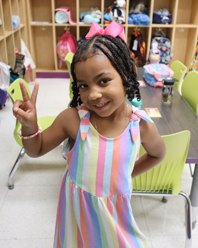 A preschool girl in a striped dress and pink bow smiles at the camera while making a peace sign