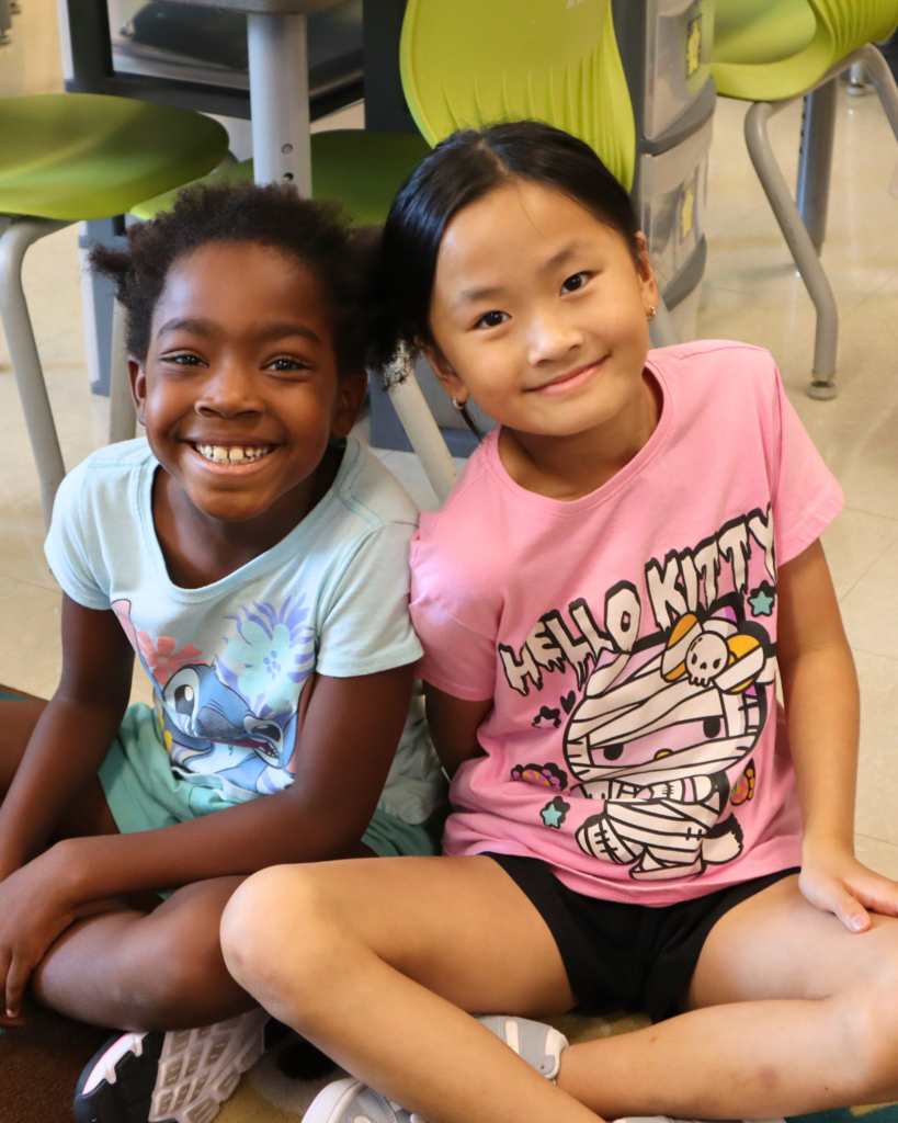 Two elementary girls sit criss cross on the carpet while smiling at the camera
