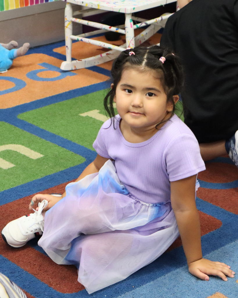 A preschooler sits on the carpet with her class in a purple dress with white sneakers and dark hair. She's looking at the camera and smiling.