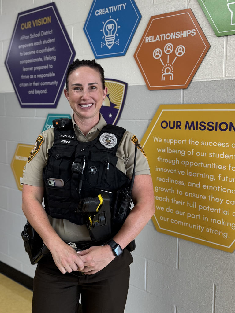Officer Ashley Meyer, wearing her police uniform and vest, smiles while standing in front of a colorful school wall display with hexagon signs that highlight Affton School District’s vision, mission, and values.