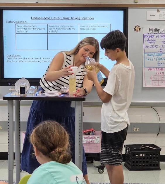 A teacher and student smiling as they pour liquid into a bottle during the homemade lava lamp experiment, with classmates watching.