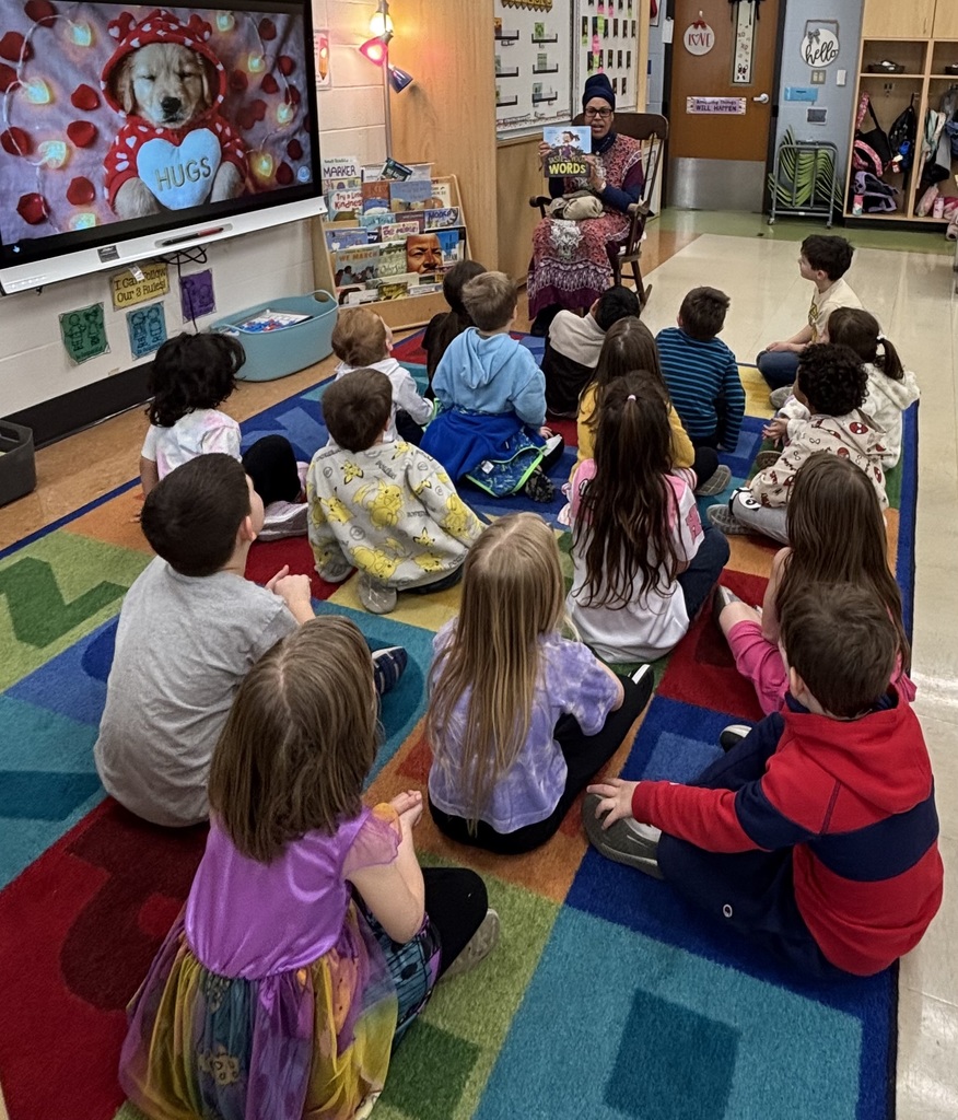 A wide, high-angle shot of a colorful kindergarten classroom. Dr. Suggs sits at the front in a wooden rocking chair, holding up a book for a class of approximately 15 students who are seated on a bright, multi-colored alphabet rug. To the left, a large digital screen displays an image of a puppy wearing a heart-themed hoodie with the word "HUGS." The room is filled with educational posters, a bookshelf stocked with diverse titles, and student cubbies in the background.