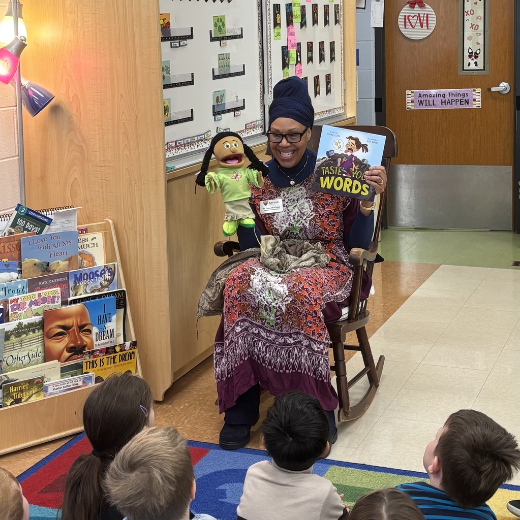 A close-up, eye-level shot of Dr. Cassandra Suggs, Assistant Superintendent, sitting in a wooden rocking chair in a kindergarten classroom. She is smiling broadly while reading the book "Taste Your Words" to a group of students. She is wearing a vibrant, patterned purple and orange African robe with a matching dark navy headwrap. In her right hand, she holds a puppet with long black braids, and in her left, she holds the book open toward the children. Several children are visible in the foreground with their backs to the camera, looking up at her attentively.