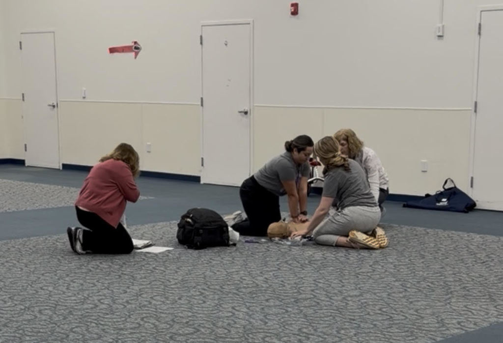 Four people in a large room practice CPR on a medical mannequin laid on the floor during a training session.