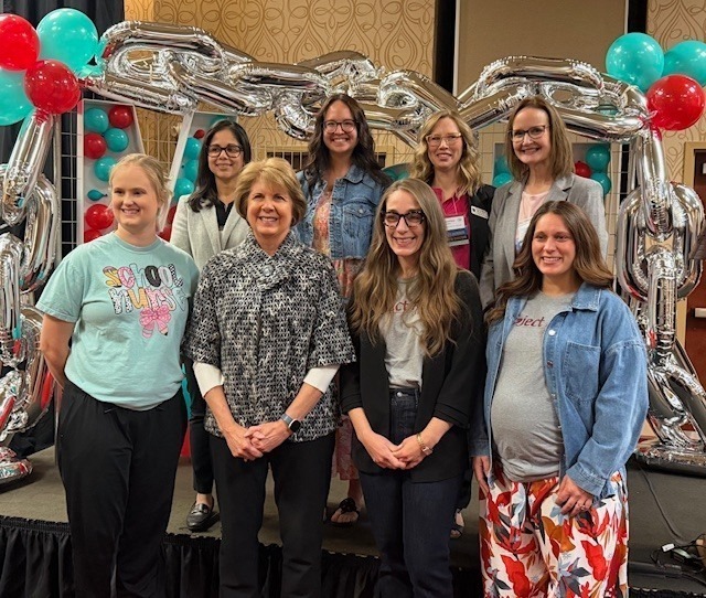 A group of seven smiling women poses in front of a decorative silver chain-link balloon arch with red and teal accents.
