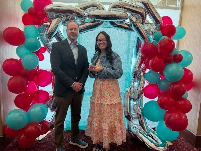 A man in a blazer and a woman in a floral dress holding a star-shaped award pose under a silver chain balloon arch.