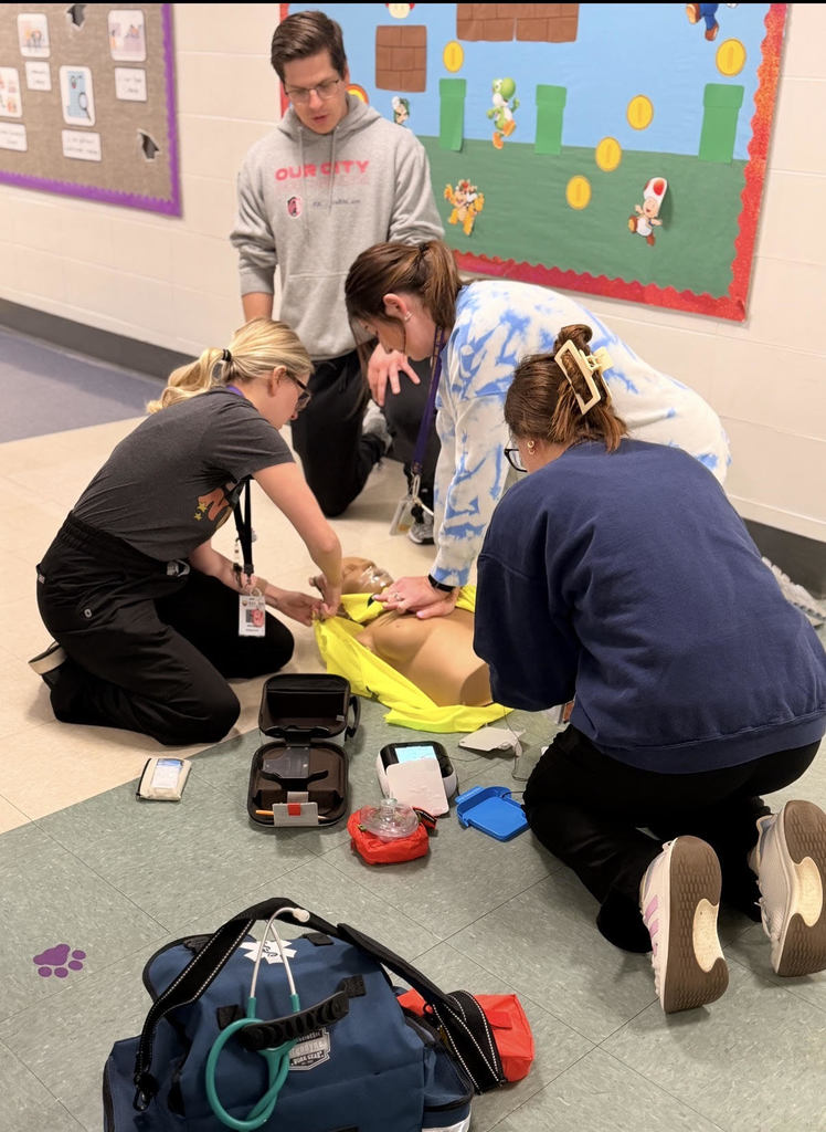 Four adults in a school hallway practice life-saving skills on a medical mannequin using an AED and first aid kit.