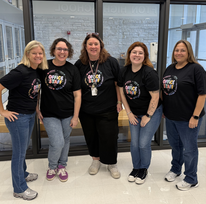 Five female school office staff members standing in a line wearing matching black "OFFICE STAFF" t-shirts and jeans.