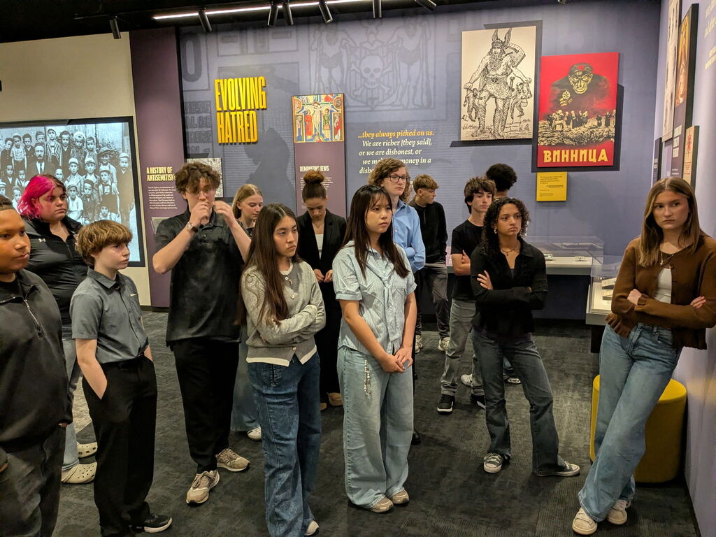Students listen attentively while standing in a museum gallery featuring historical Holocaust exhibits.