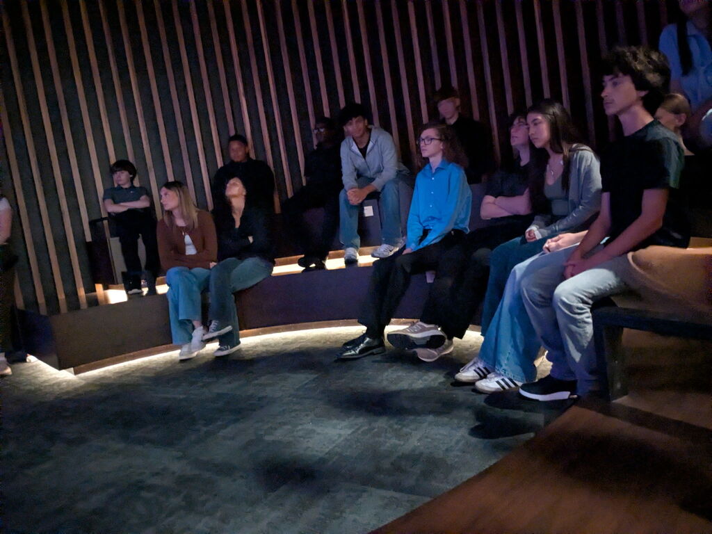 Students sit on tiered wooden benches in a dimly lit museum theater watching a presentation.