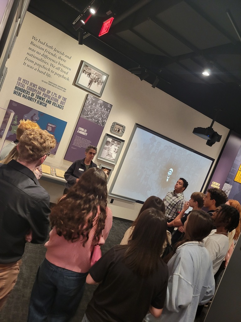 A guide speaks to a group of students gathered around historical wall displays and photos.
