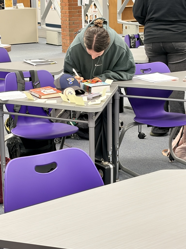 Person with a bun in a green hoodie leans over a table, writing on paper next to a baseball cap and notes.