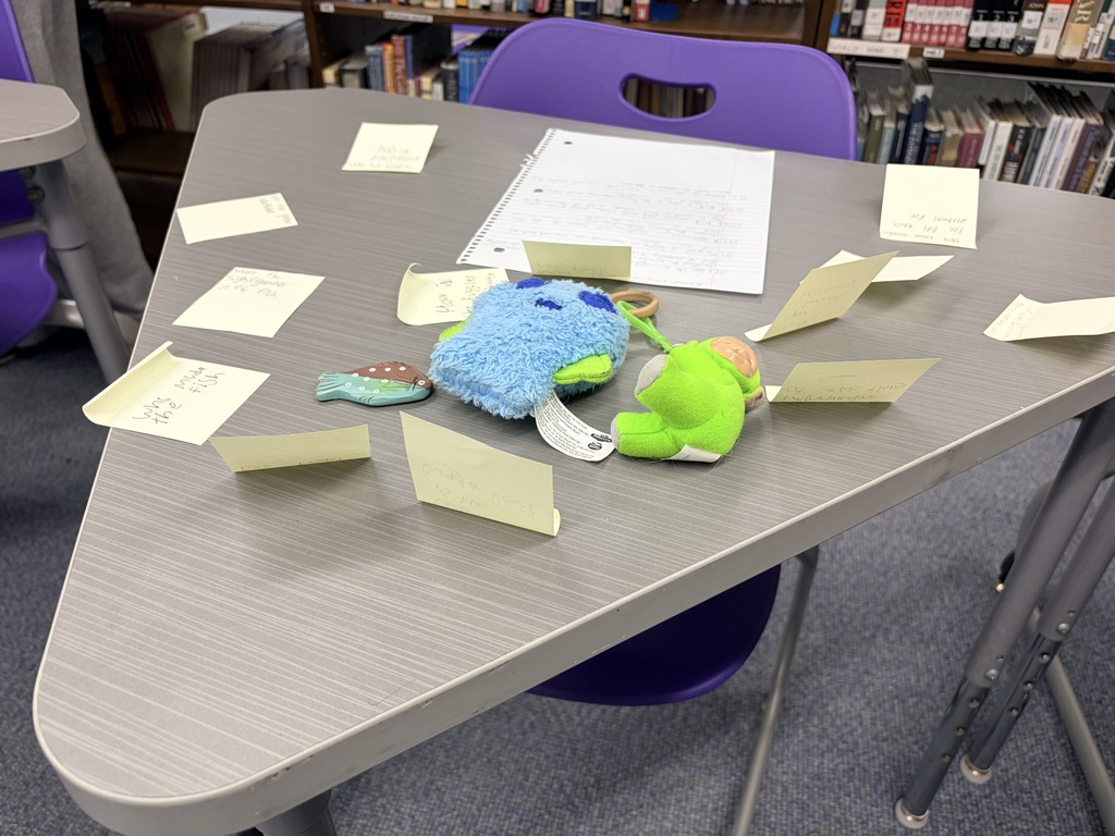 Blue plush, green doll, and notepad on a desk with many fan-shaped sticky notes in a library.