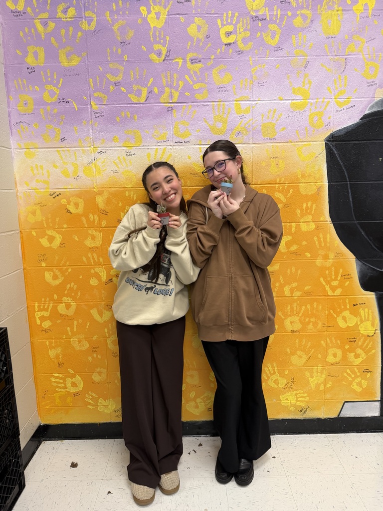 Two smiling students stand together holding small potted succulent plants in front of a bright yellow handprint wall.