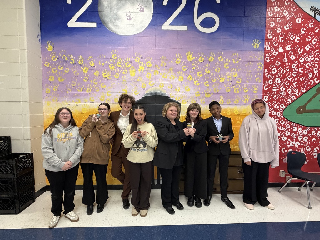 A group of seven students stand together smiling and holding small potted succulent plants in front of a graduation mural.