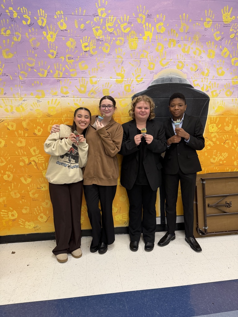 Four smiling students stand side by side holding small potted succulent plants against a colorful handprint mural.
