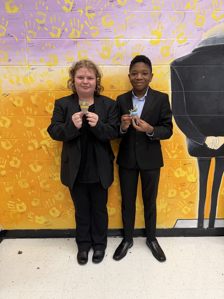 Two students wearing black suits pose together while holding small potted succulents in front of a yellow handprint mural.
