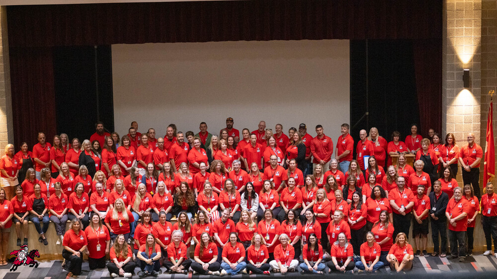 All Afton Central School District teachers, faculty and staff gather for a group photo on an auditorium stage, each wearing a red shirt with the Afton Crimson Knights logo