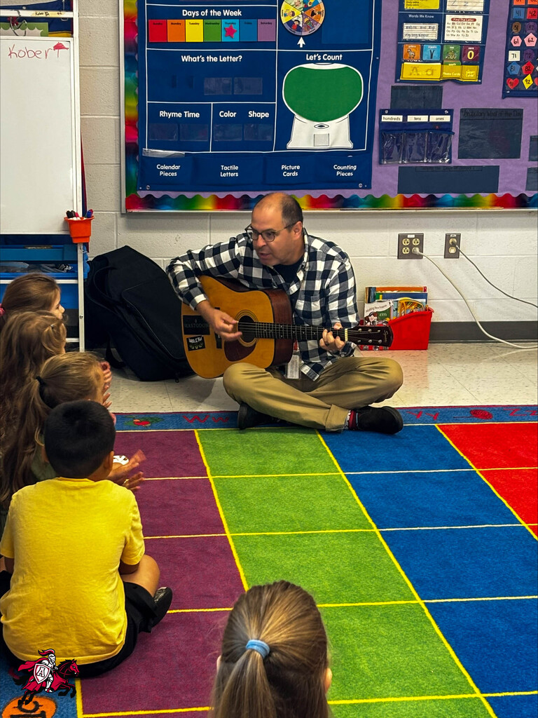 A teacher plays the guitar as a group of pre-K and kindergarten students gather around and listen