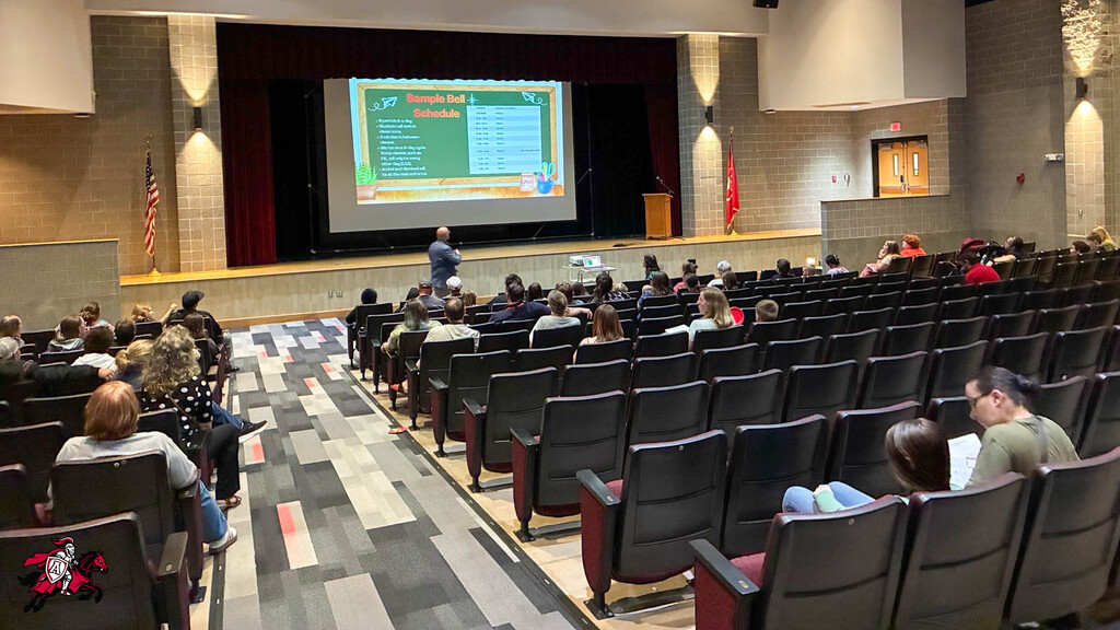 Middle/High School Principal Joel Carle stands in front of a large projector screen at the front of an auditorium to share important information with 6th graders and their parents who are watching and listening from the auditorium chairs