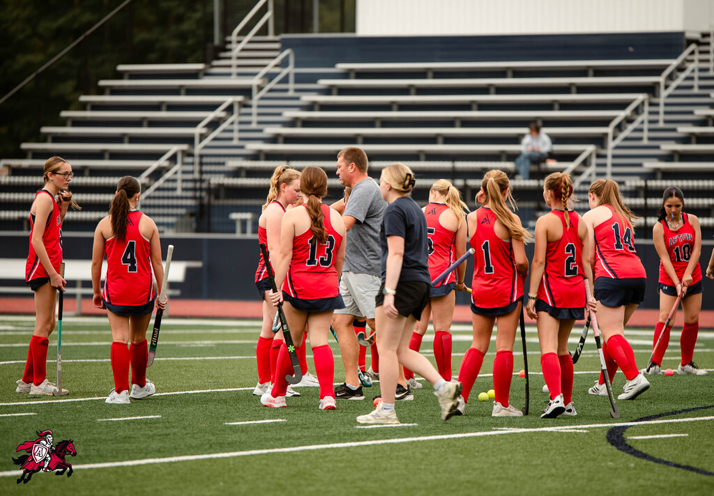 A group of female players in red uniforms and shorts gather on a field, holding hockey sticks.