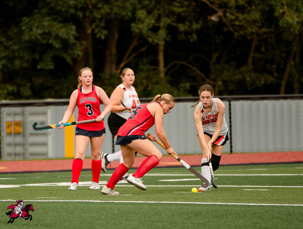 Four women in sports uniforms play field hockey on a field. One woman bends over, and three others stand. Trees and a fence are in the background.
